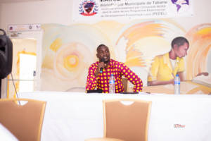 Mr. Madochée Placide sitting behind a table with a mic in his hand. There are two chairs in front of the table
