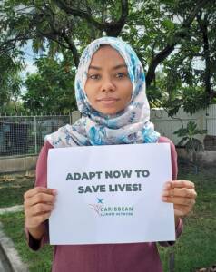 Earth Day photo petition by CCN Member in Guyana. A young woman is holding a sign which reads 'Adapt Now to Save Lives'.